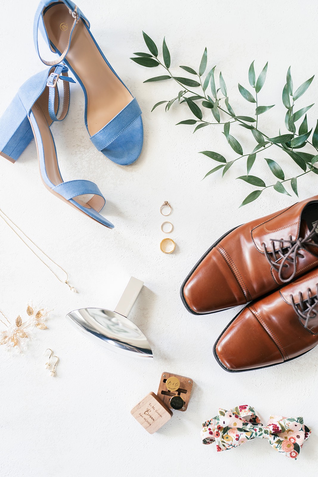 Flat lay of blue bridal heels, brown groom's shoes, rings and floral bow tie