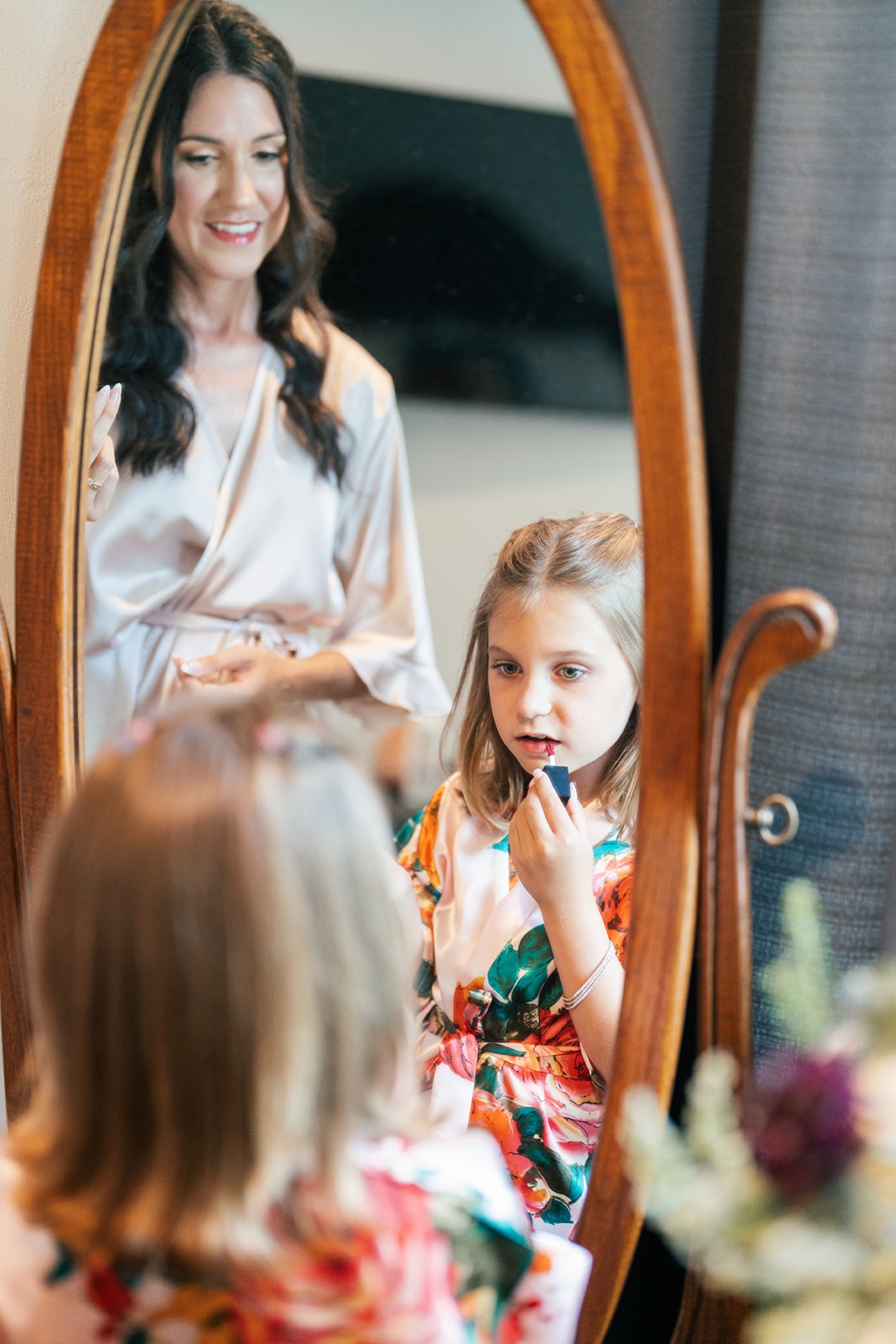 Young flower girl applies lipstick at a mirror while Danielle looks on in her robe