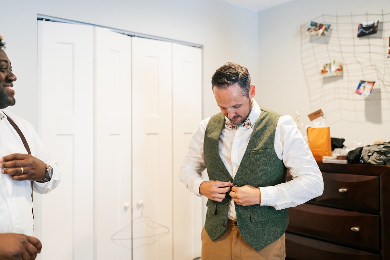Brian buttons his green vest and bow tie while a groomsman waits nearby