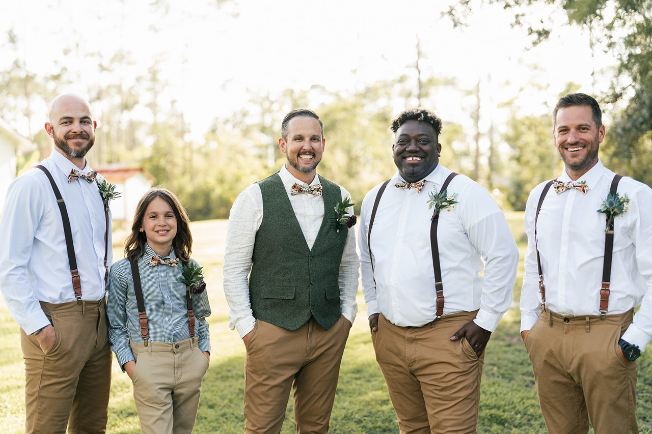 Brian stands with his groomsmen in suspenders and bow ties on a grassy lawn