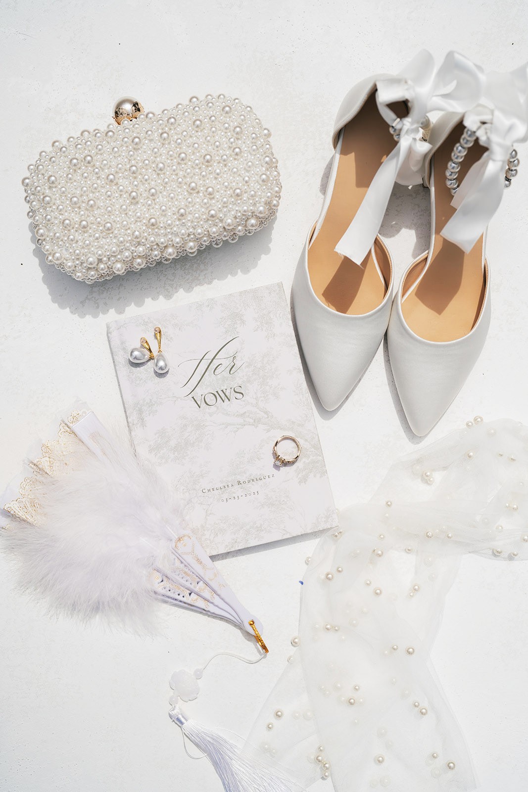 Bridal details flat lay with pearl clutch, white heels, vow book, earrings and ring