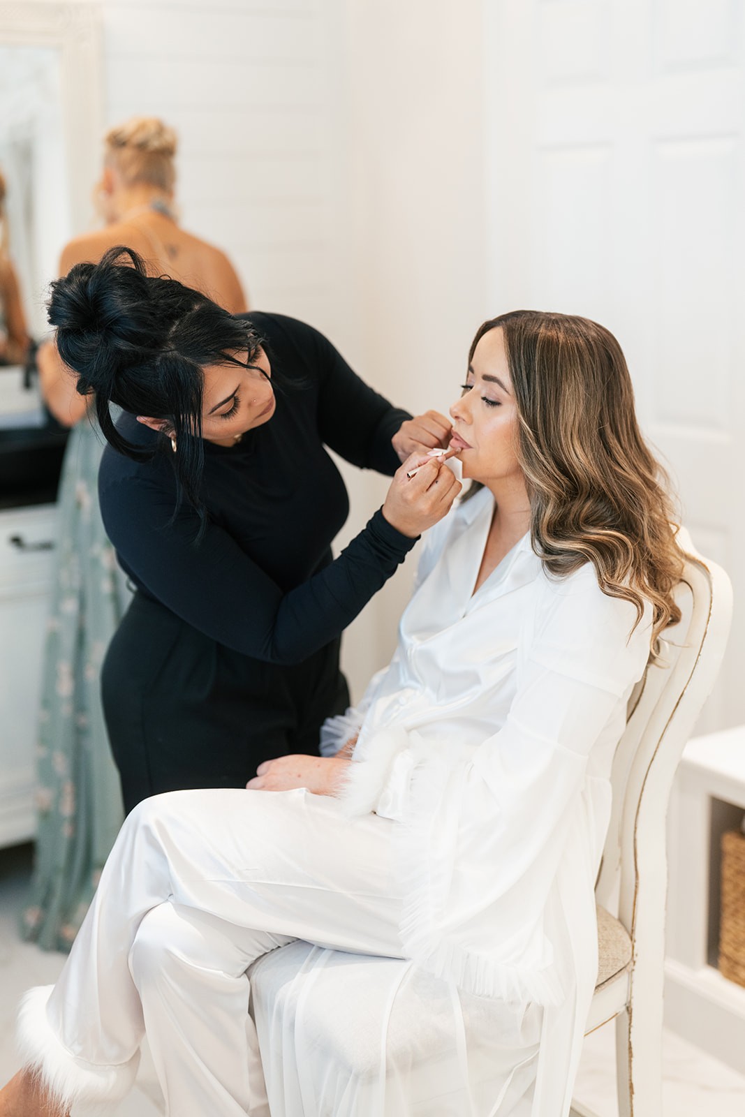 Makeup artist applying lipstick to Chellsea seated in a white robe before the wedding