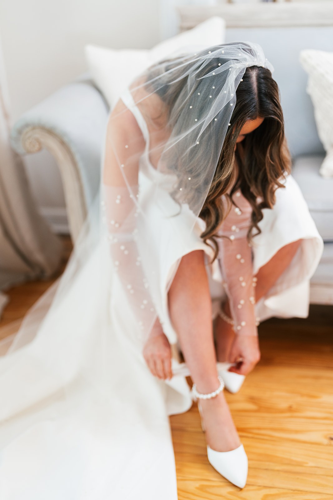 Chellsea in pearl veil bending to fasten her white ankle-strap heels