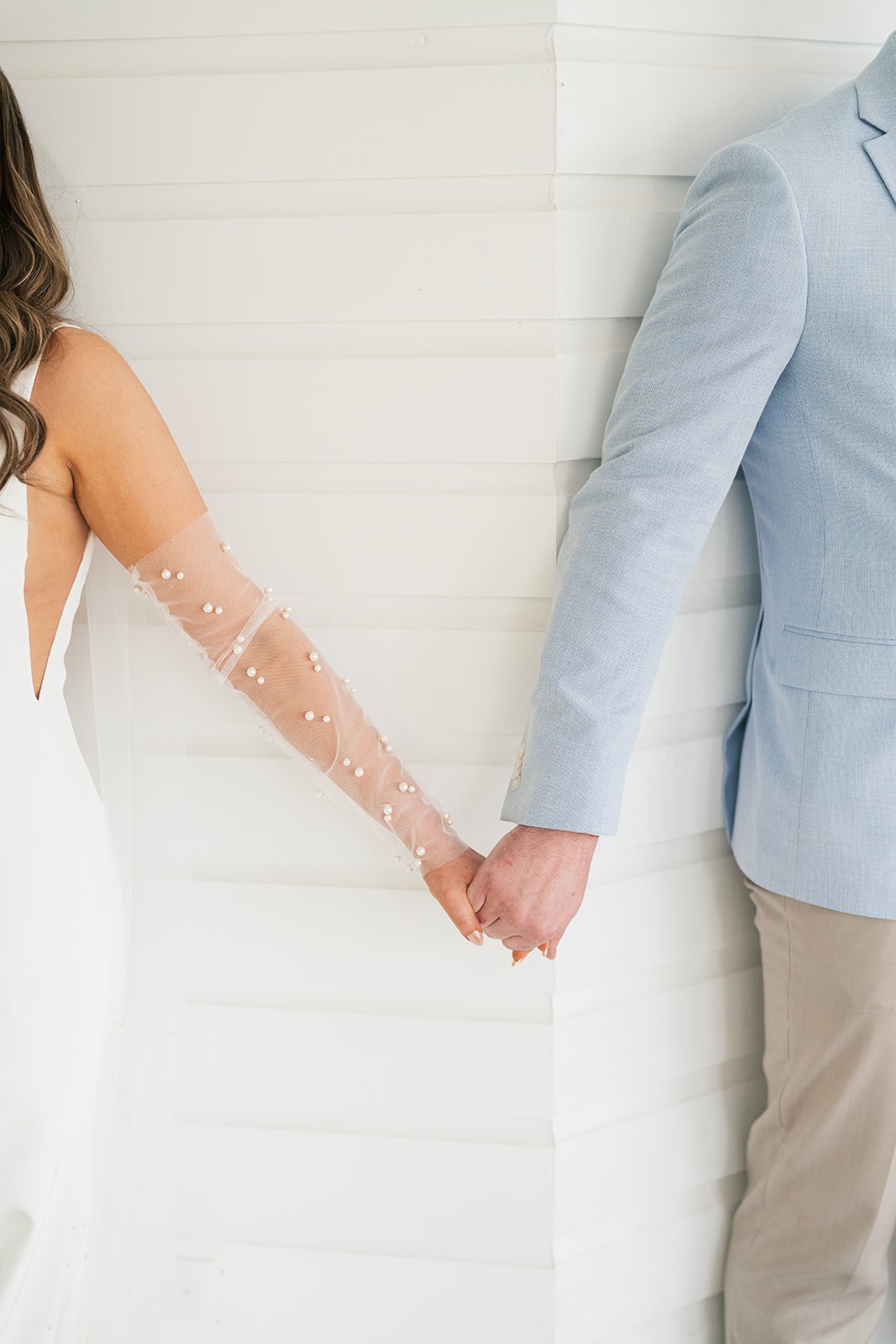 Chellsea and Jacob holding hands against a white wall, her pearl sleeve catching the light