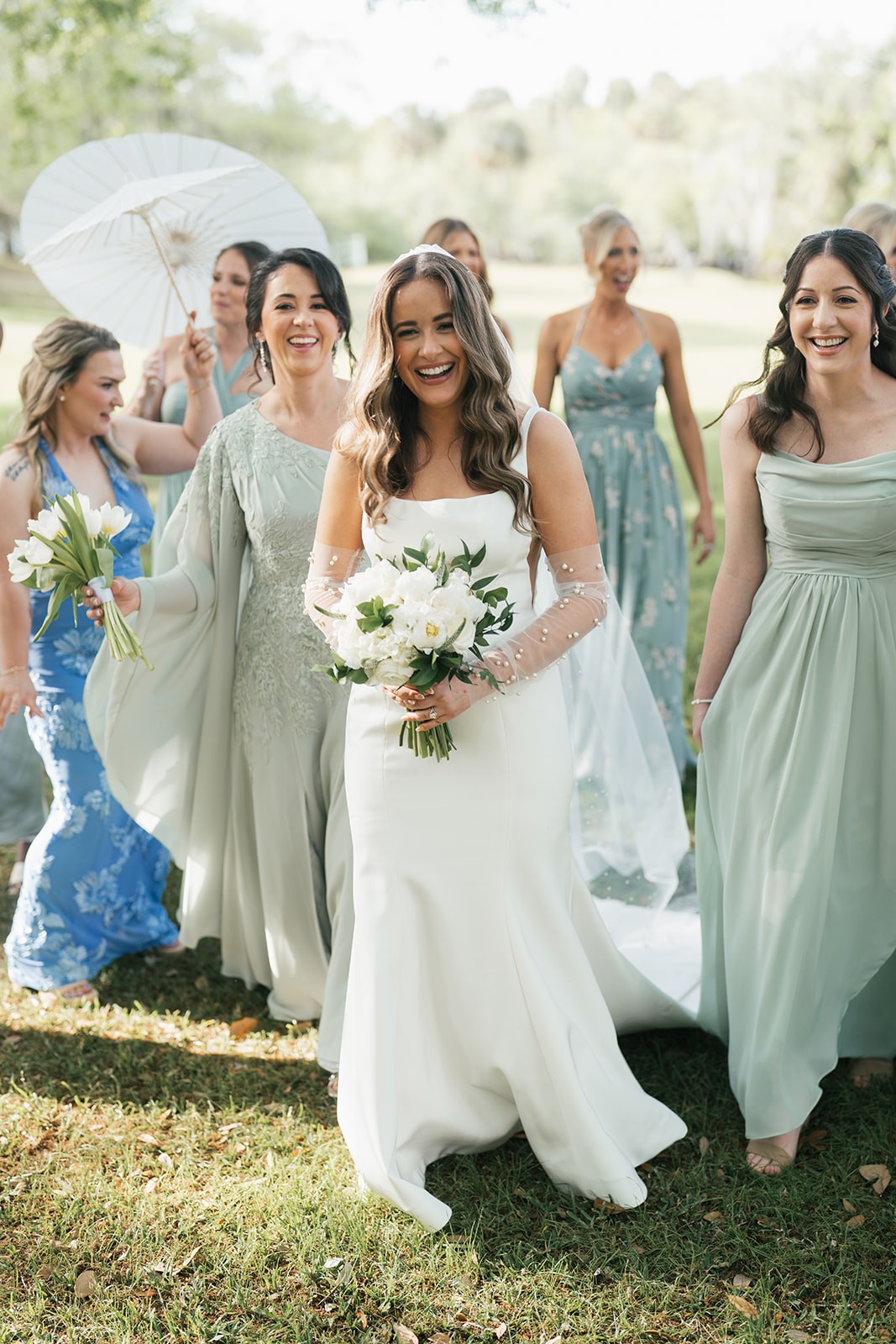 Chellsea walking with her bridesmaids in sage gowns, holding a white bouquet on the lawn