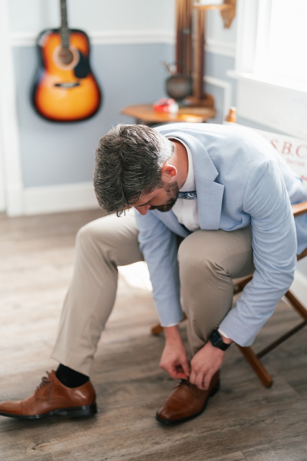 Jacob leaning forward to tie his brown dress shoes near a guitar on the wall
