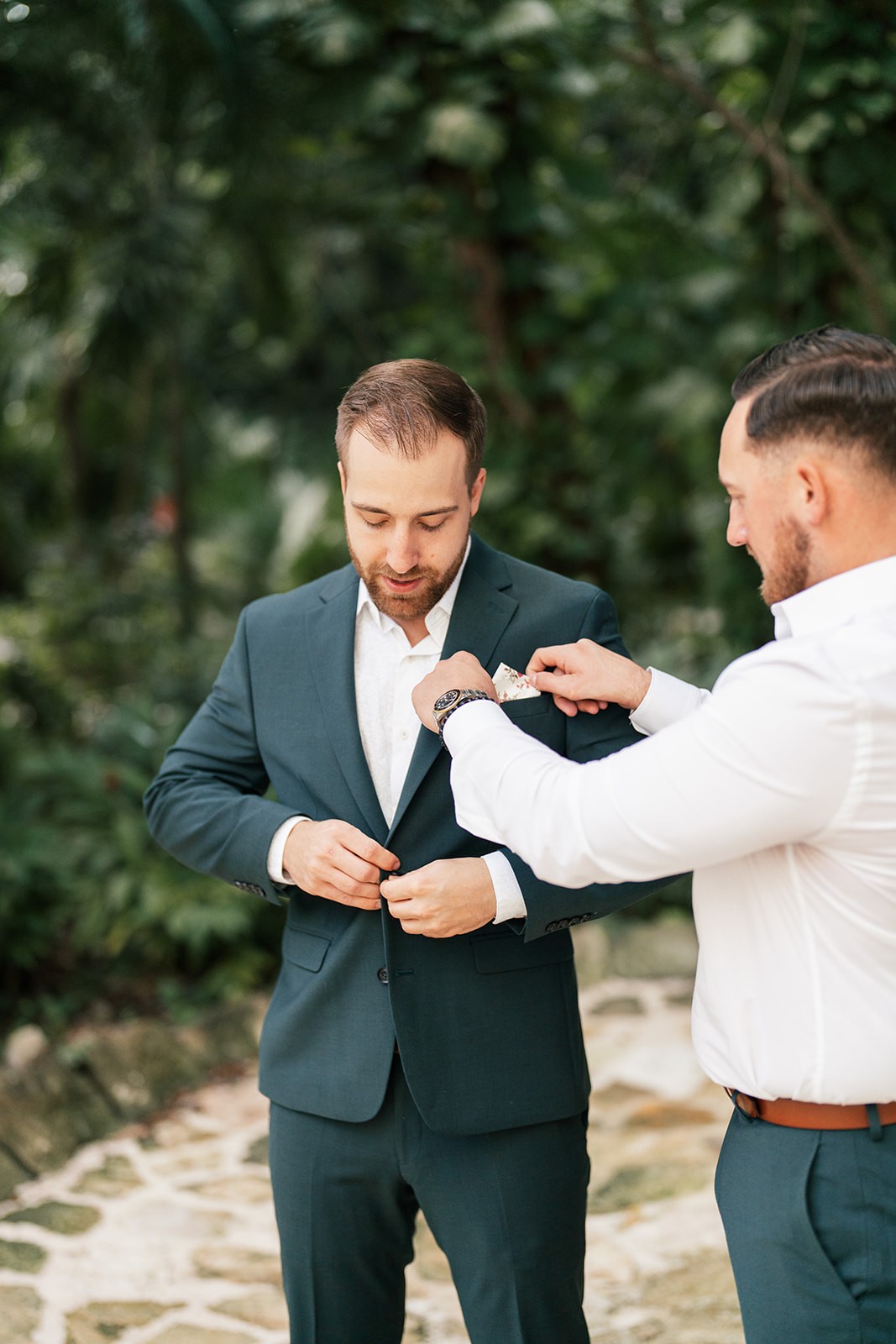 Groomsman helps Brian button his dark suit jacket on a garden stone path