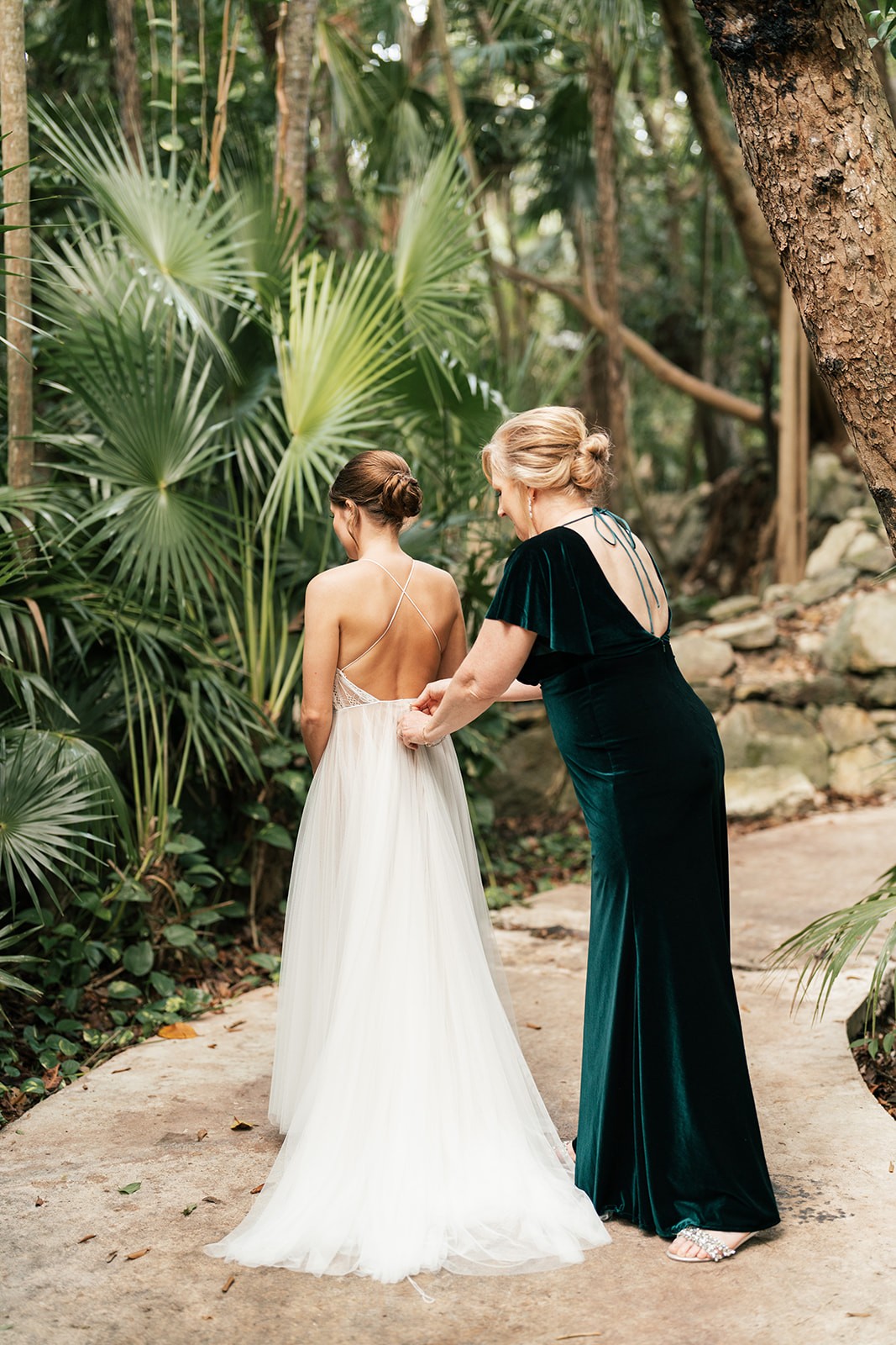 Woman helps button Lindsey's open-back gown on a stone path among lush palms