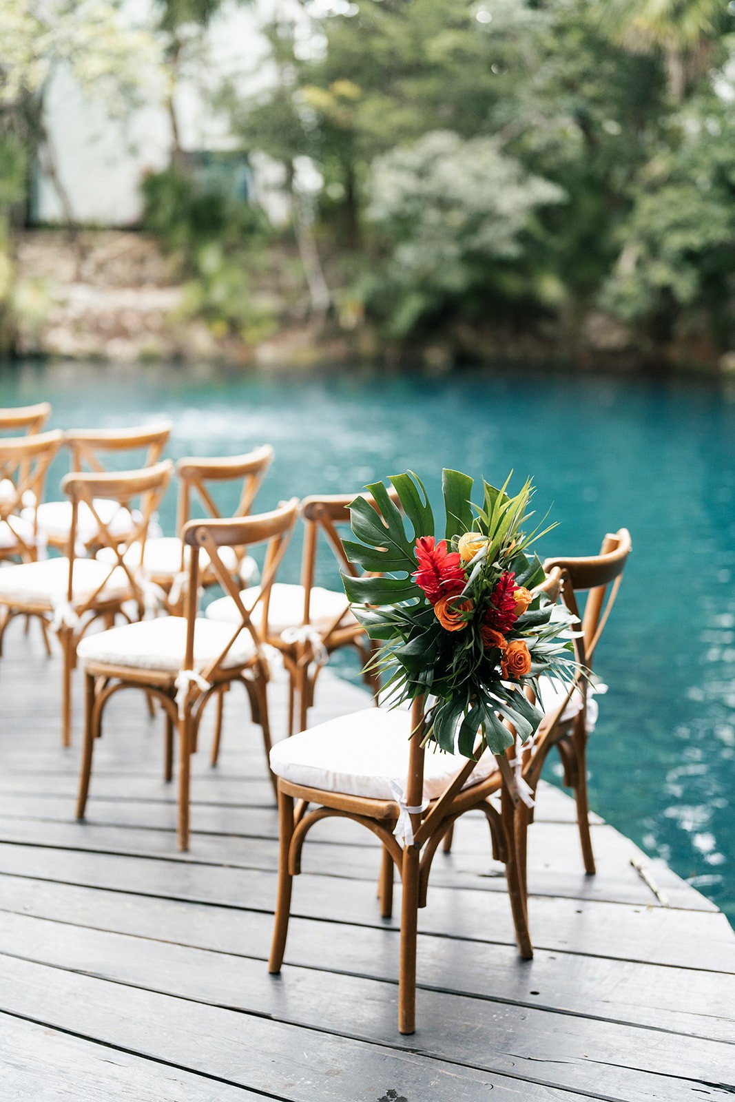 Wooden ceremony chairs on a dock by turquoise water, one with a tropical bouquet