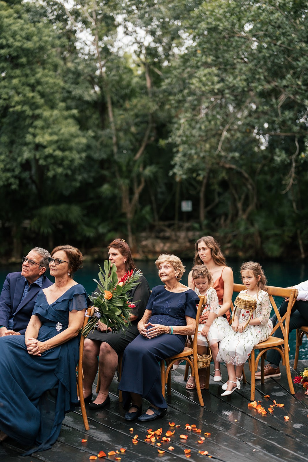 Family guests seated on wooden chairs watching the ceremony, petals scattered on the dock