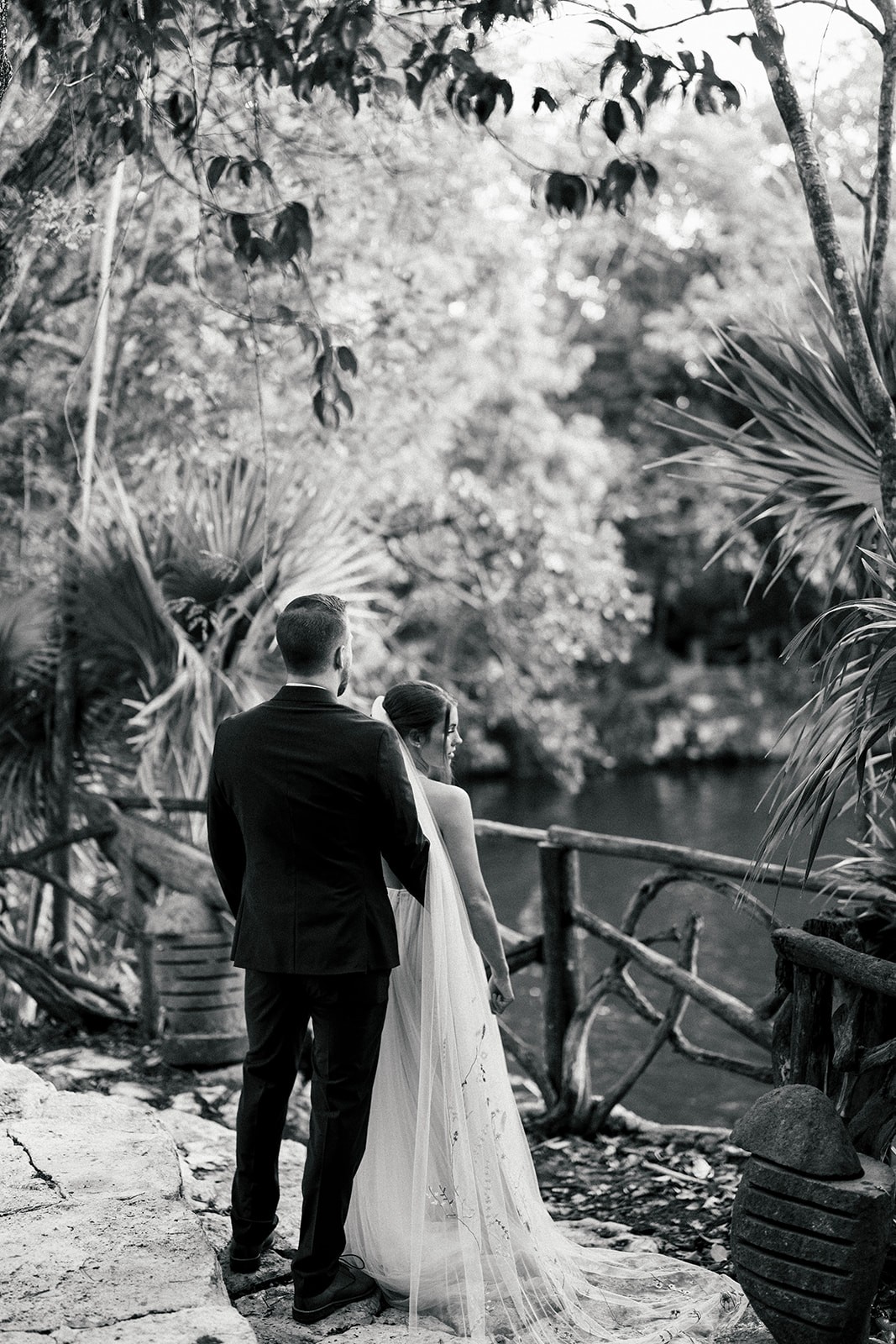 Black and white of the couple overlooking water from a railing in dense foliage