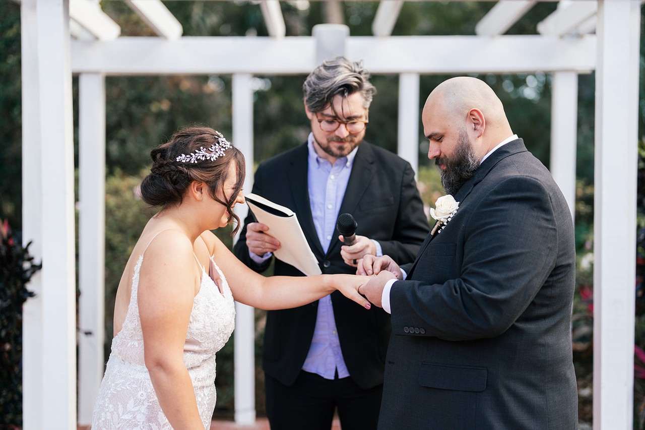 Manny placing the ring on Sydney's hand during their outdoor ceremony beneath a white arbor