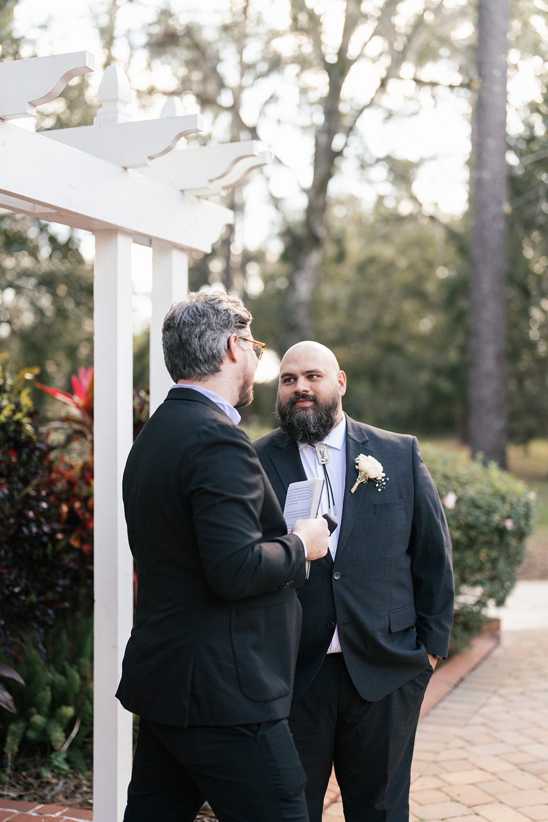 Manny speaking with the officiant beside a white arbor in a leafy garden before the ceremony