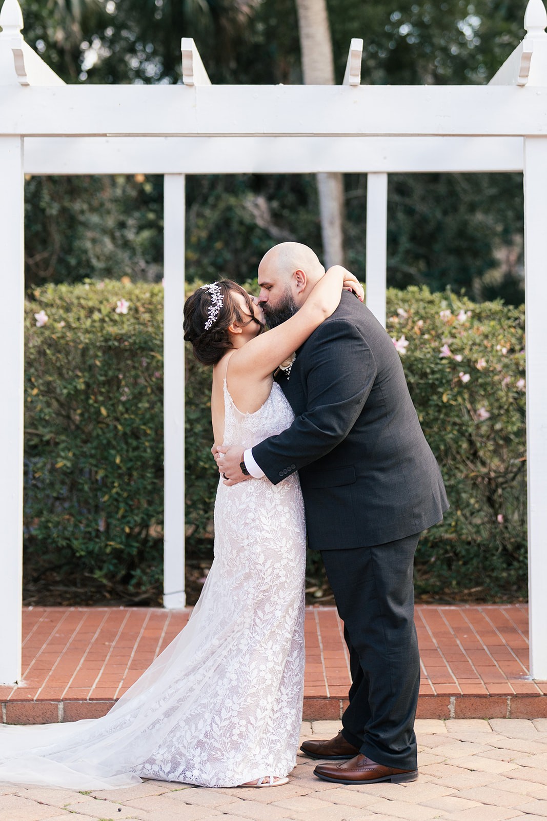 Sydney and Manny embracing in front of a white pergola and green hedges on a brick patio