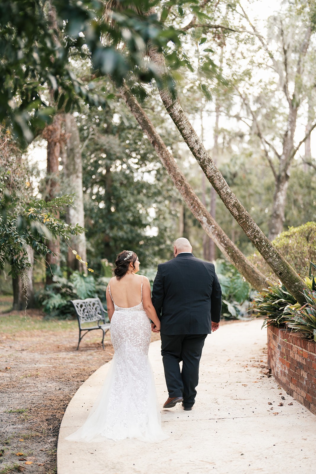 Sydney and Manny walking hand in hand down a tree-lined garden path away from the camera