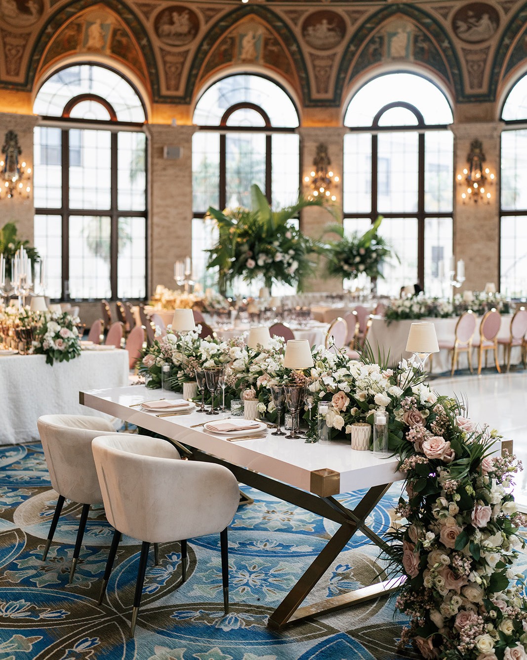 Sweetheart table dressed with roses and taper lamps beneath the frescoed arches of a grand ballroom reception