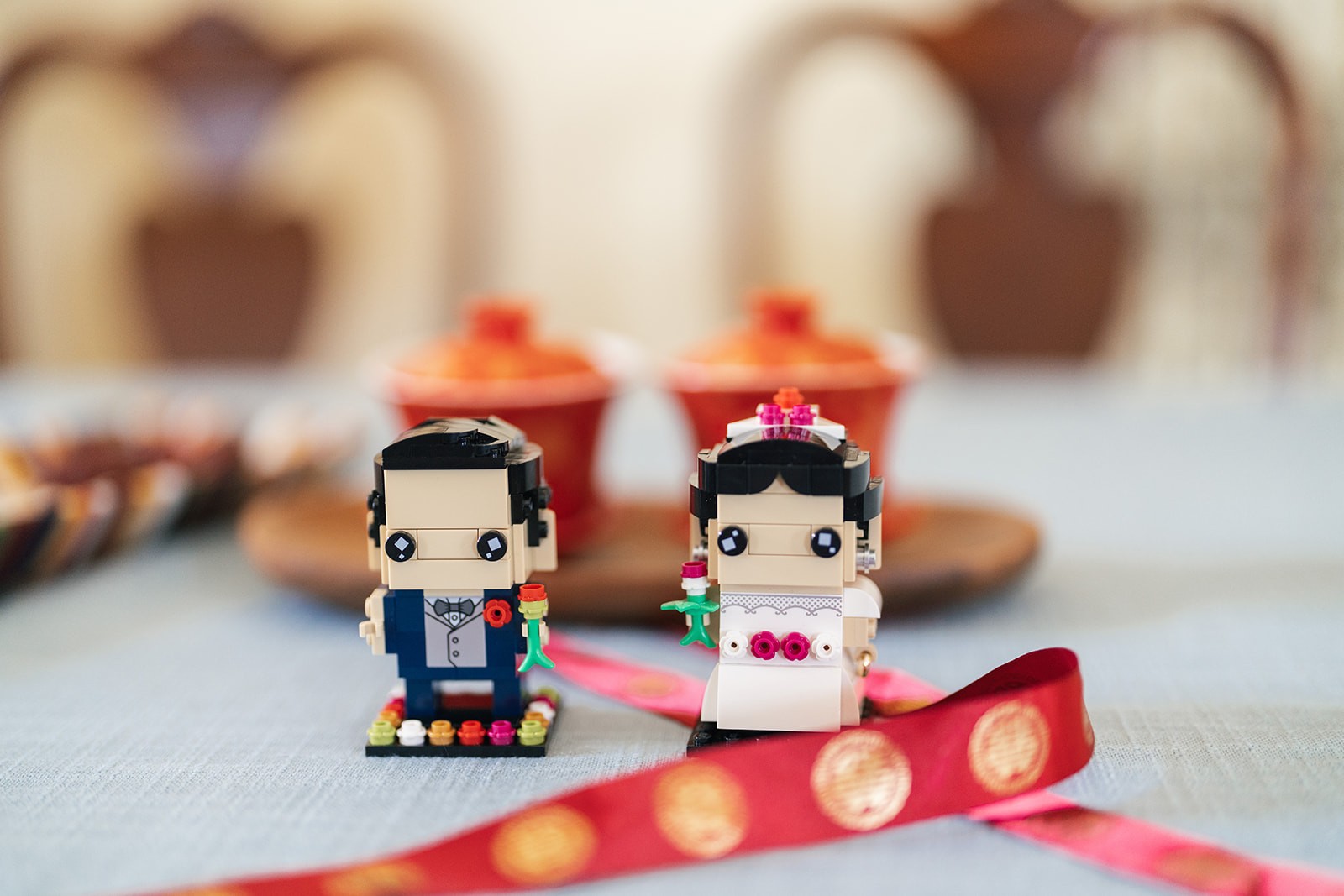 LEGO BrickHeadz bride and groom figurines posed together on a table beside red ceremonial cups and a red ribbon with gold double-happiness coins.