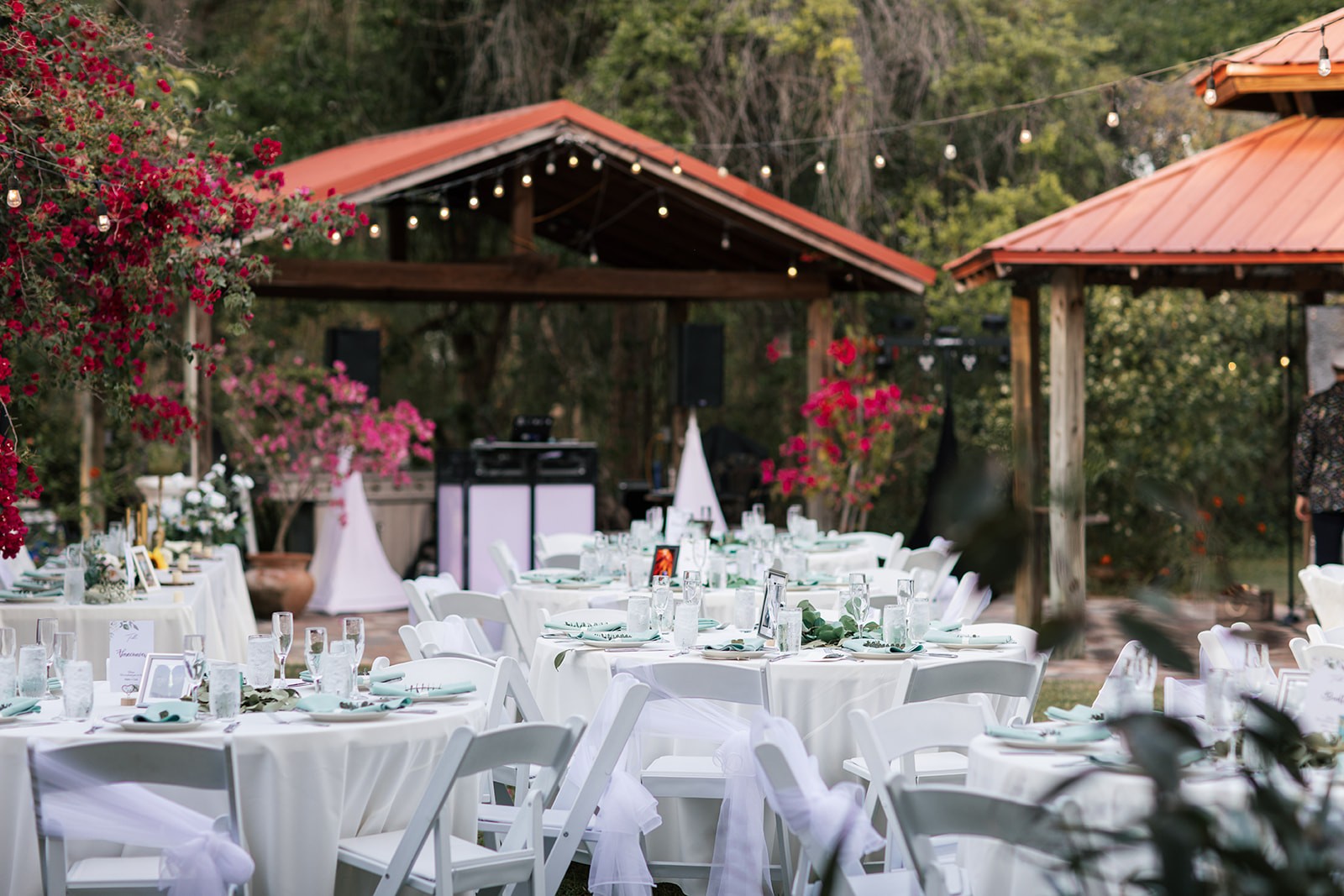 Outdoor wedding reception setup with round tables draped in white linens, sage napkins, and string lights strung between wooden gazebos surrounded by vivid bougainvillea.