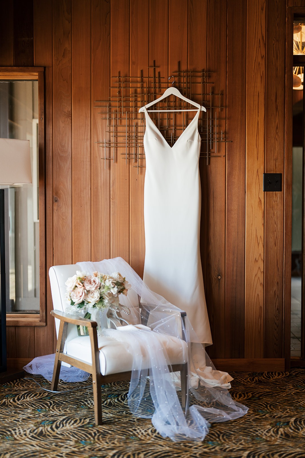 Sleek spaghetti-strap wedding gown hanging on a wood-paneled wall above a chair draped with a pearl-dotted veil and a blush bouquet.