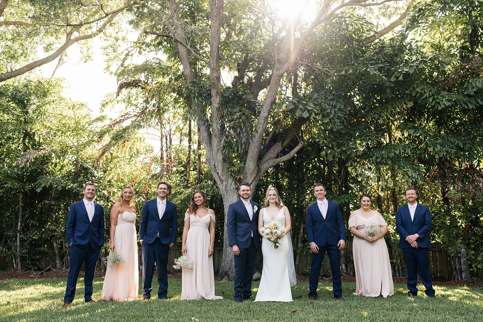 Full wedding party laughing together on a sunlit lawn, golden backlight coming through the canopy above them