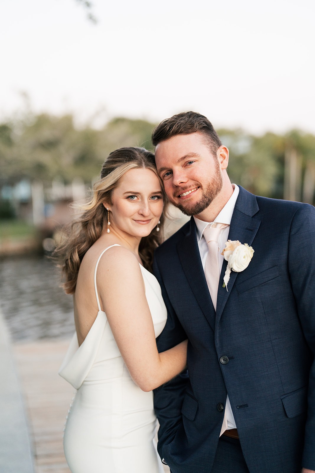 Bride and groom lean cheek to cheek smiling at the camera, waterway and palm trees softly blurred behind them.