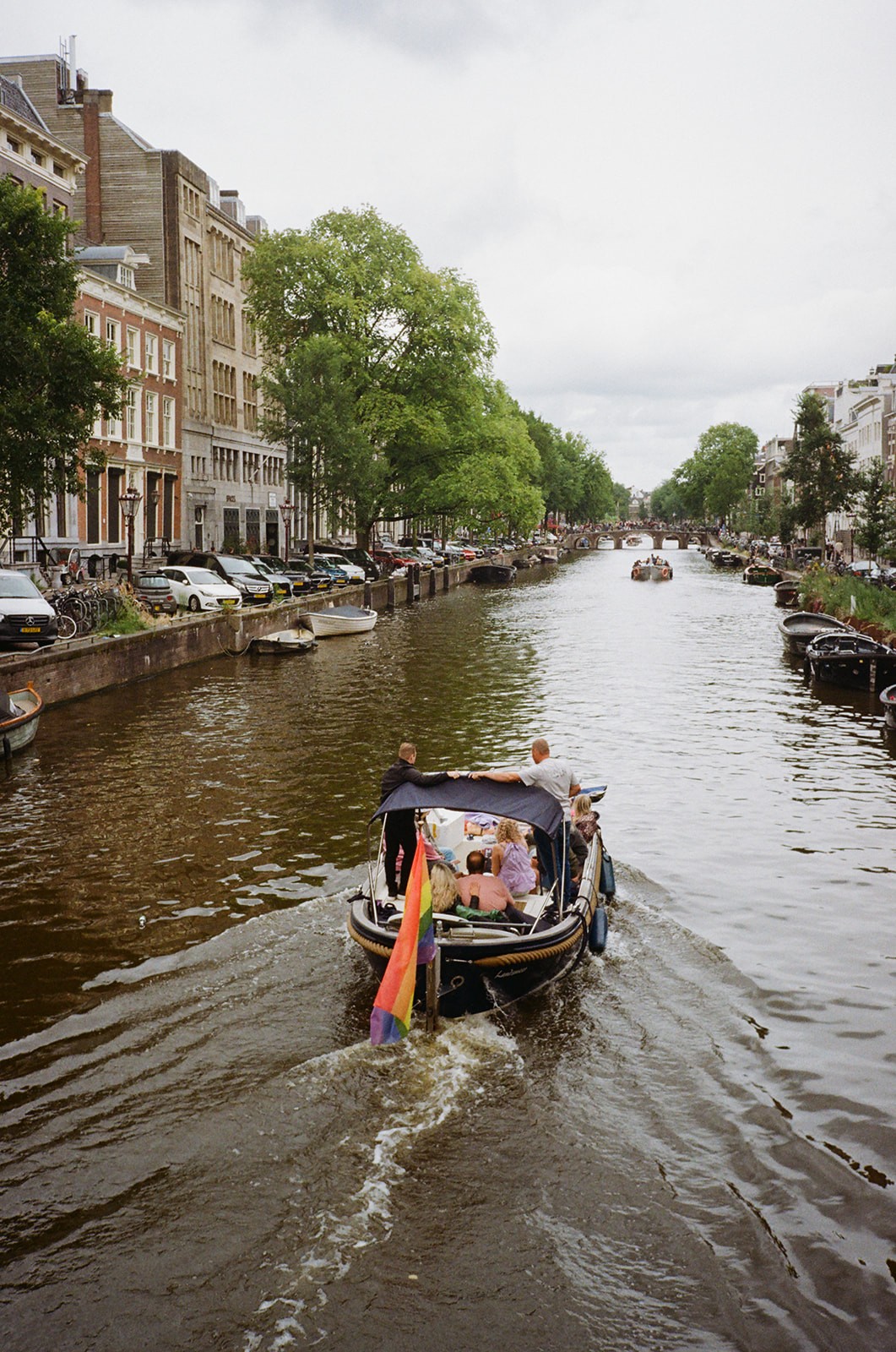 A canal boat with a rainbow flag glides past historic Amsterdam buildings and moored bicycles along the water.