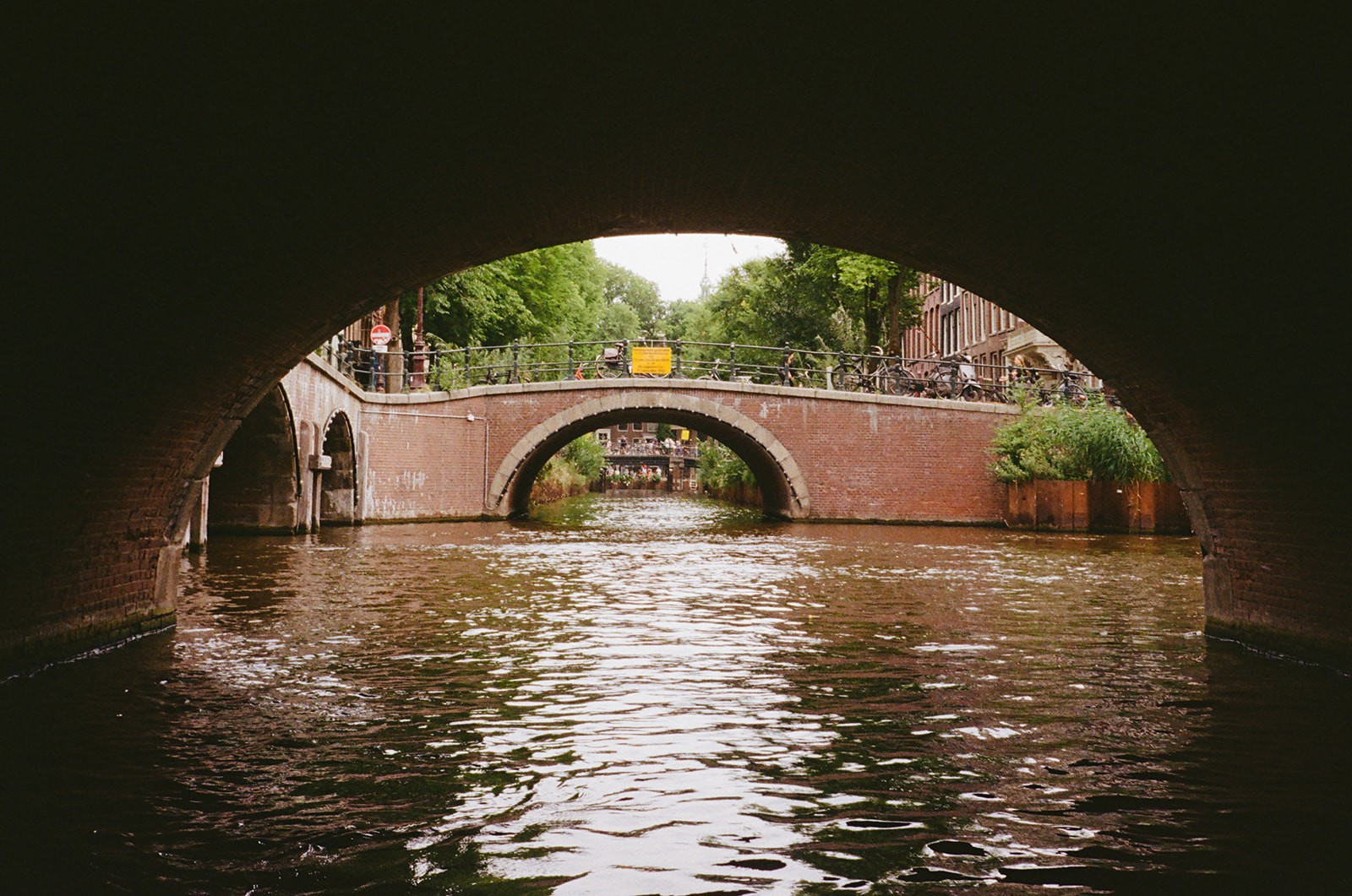 View from a canal boat passing beneath a stone bridge in Amsterdam, with a second arched bridge visible ahead.