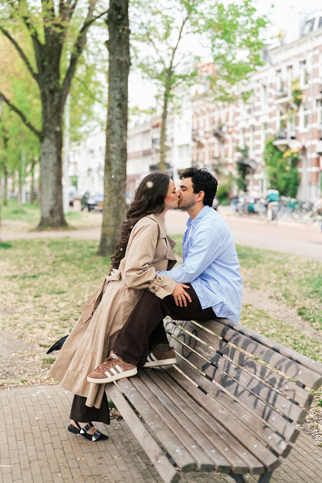 Couple kisses on a park bench along a tree-lined Amsterdam street, canal-side row houses and parked bicycles in the background.