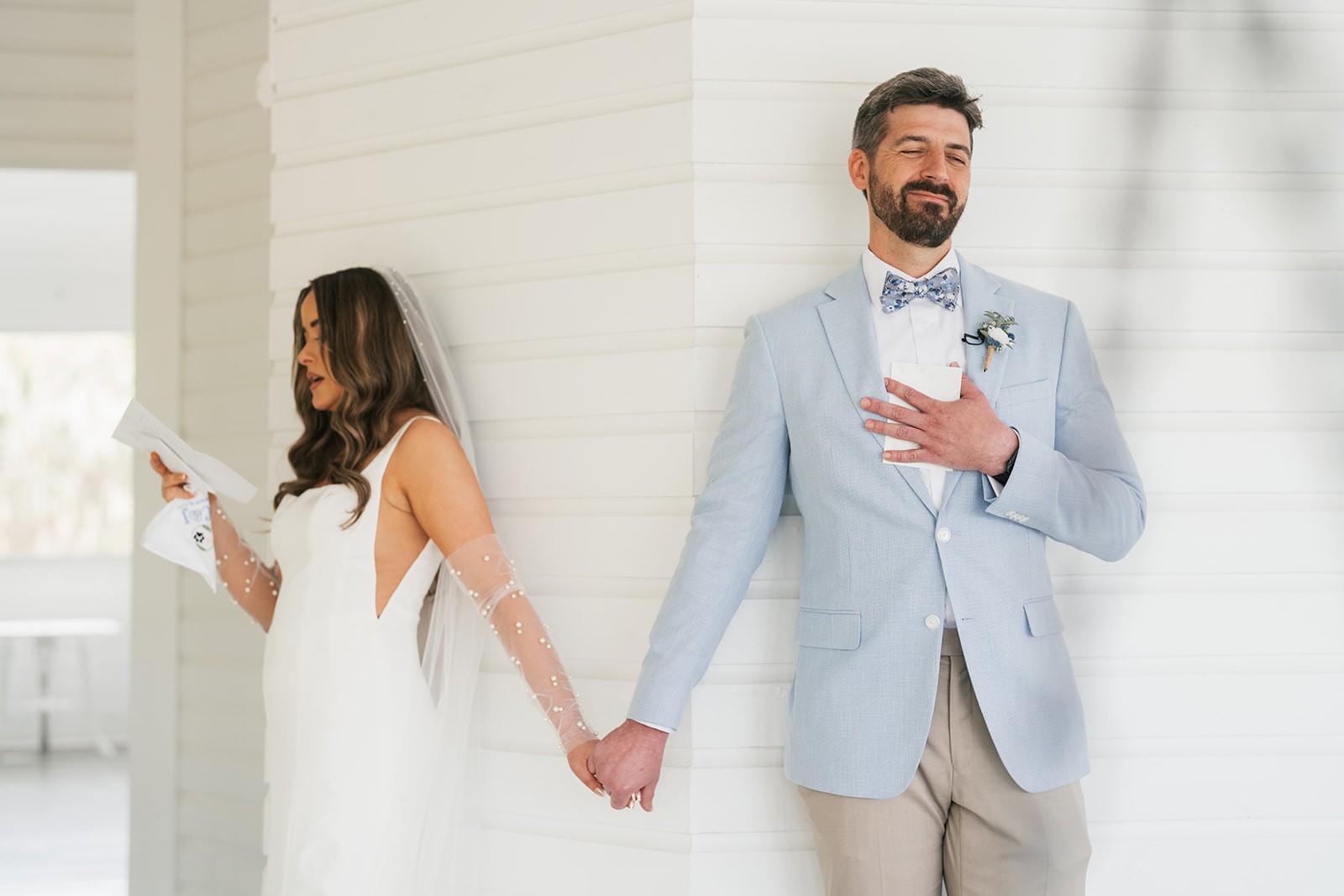 Bride and groom read their vows separately while holding hands around a white shiplap corner before their first look.
