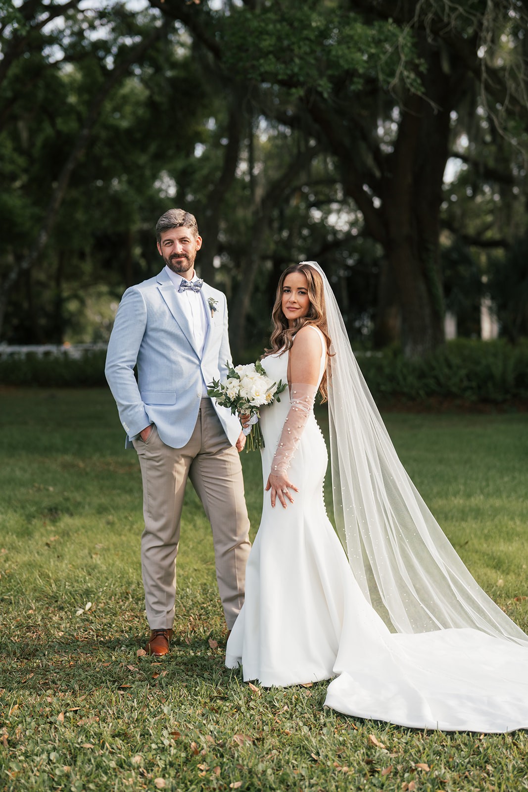 Bride in a fitted gown with a pearl-embellished veil stands beside the groom in a light blue jacket holding her white bouquet on the lawn.