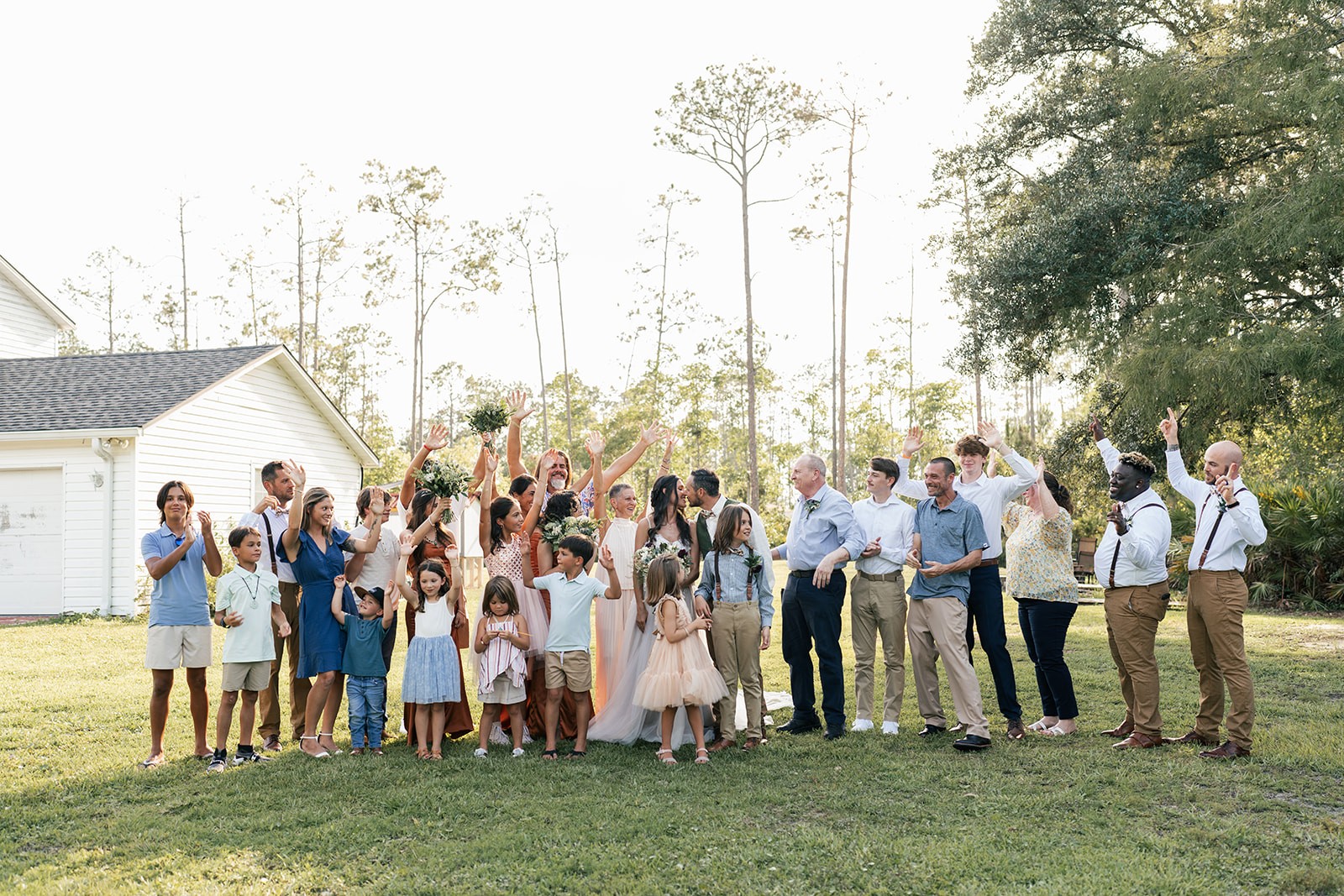 Guests cheer and toss arms in the air around the couple on the lawn at sunset