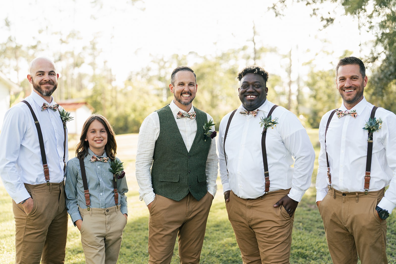 Brian stands with his groomsmen in suspenders and bow ties on a grassy lawn