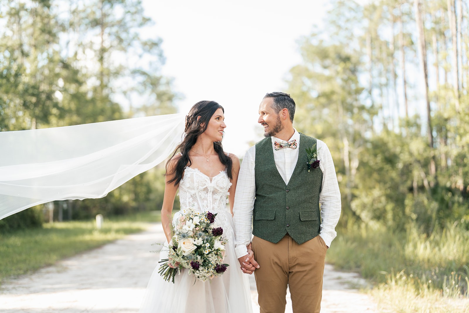 Danielle and Brian hold hands walking a sandy path, her veil drifting in the breeze