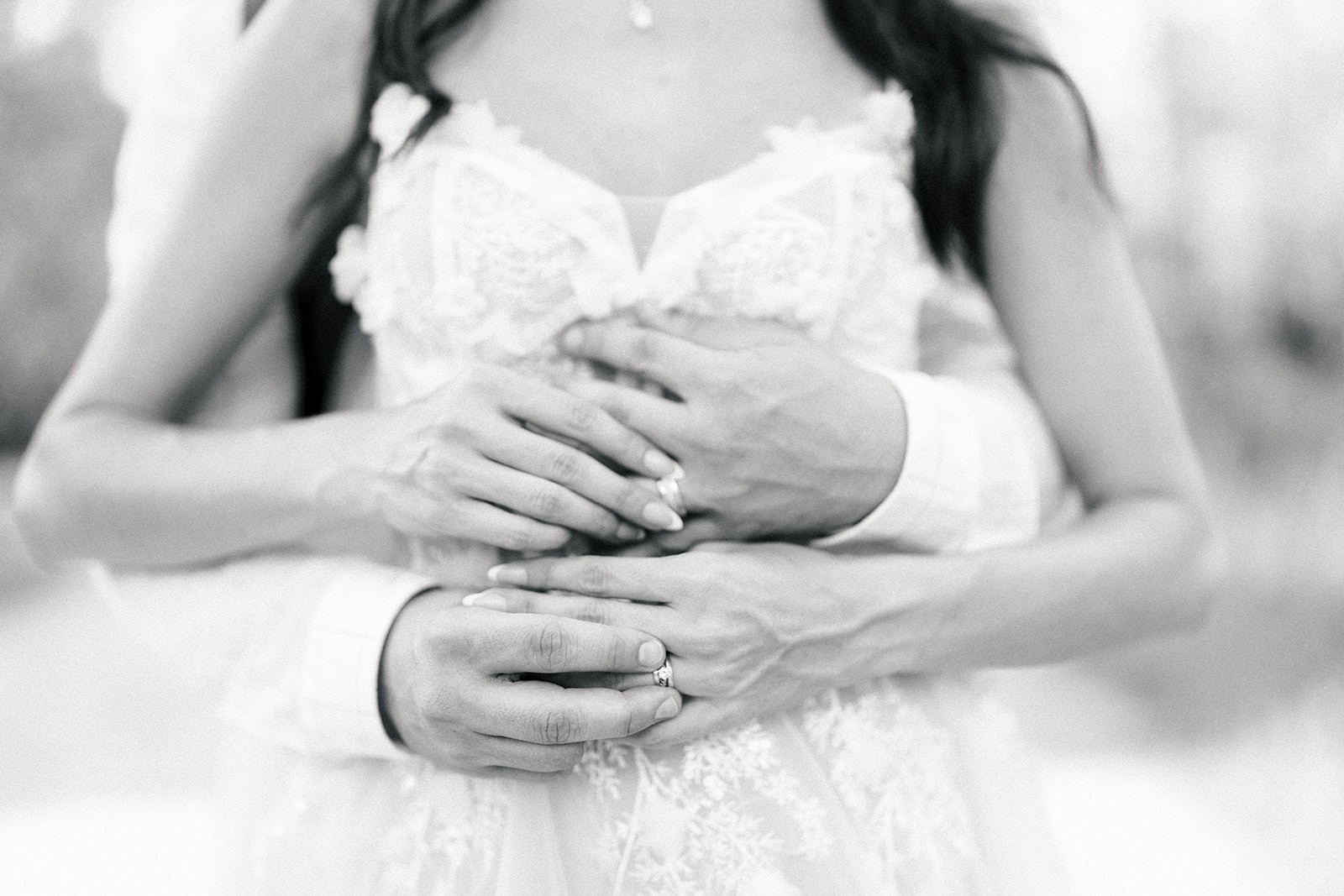 Brian's arms wrap around Danielle's waist over her lace gown, wedding rings visible