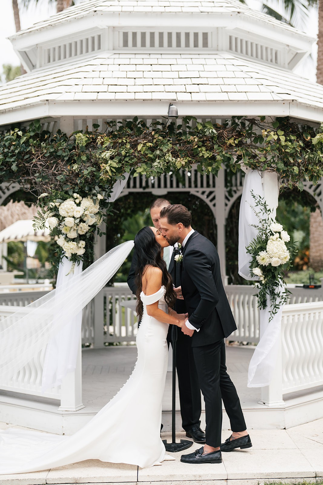 Bride and groom sharing their first kiss beneath a white gazebo adorned with white hydrangea clusters and trailing greenery, the bride's veil sweeping in the breeze