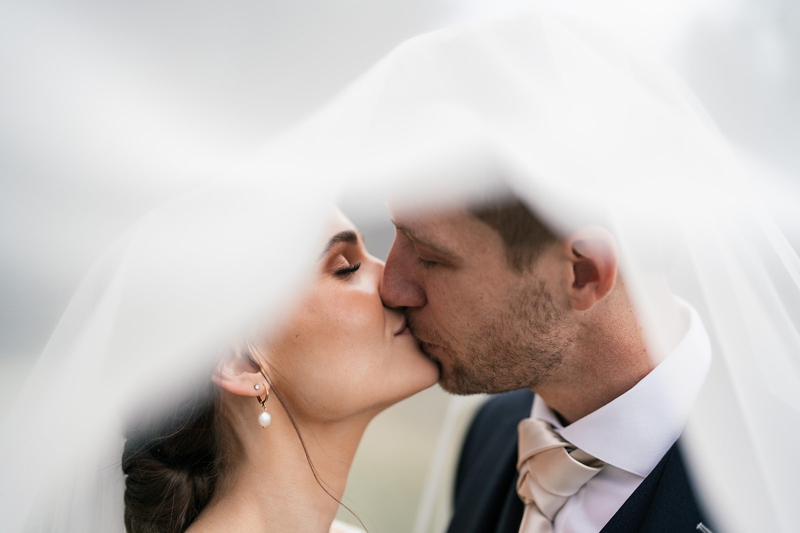 Bride and groom share a close kiss beneath a billowing veil during outdoor wedding portraits.