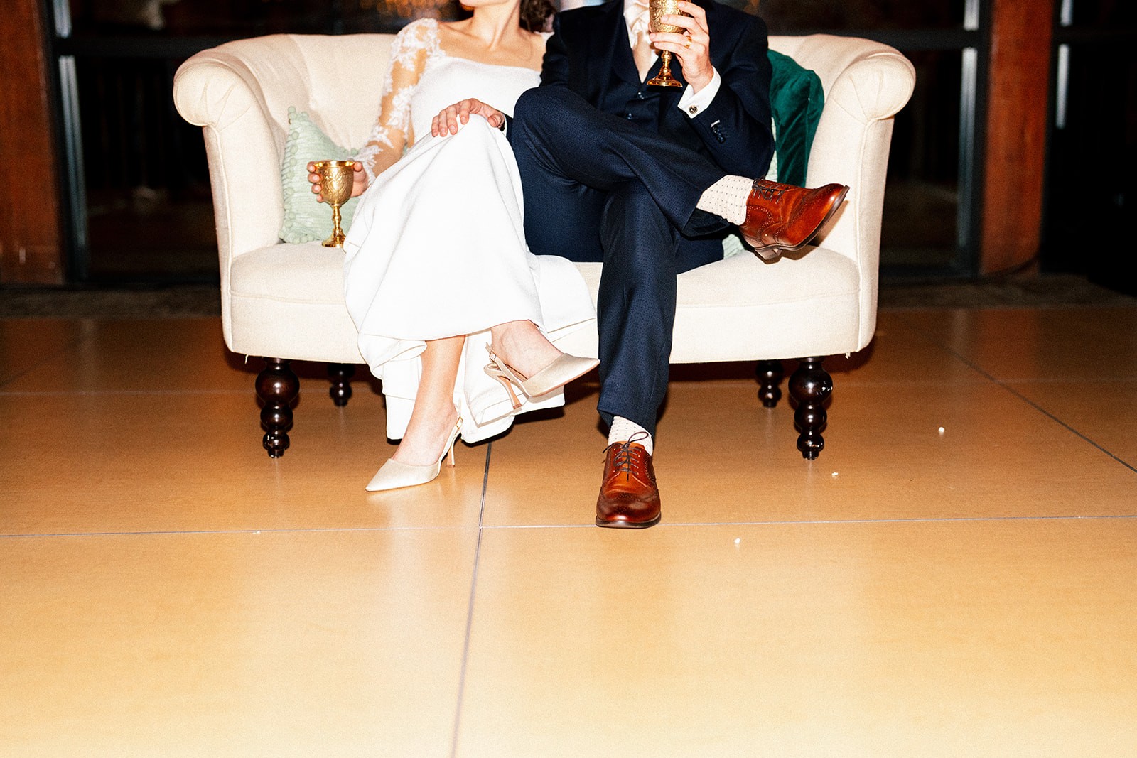 Bride and groom seated on a cream loveseat during the reception, each holding a gold chalice, cropped at the waist to show their shoes and attire.
