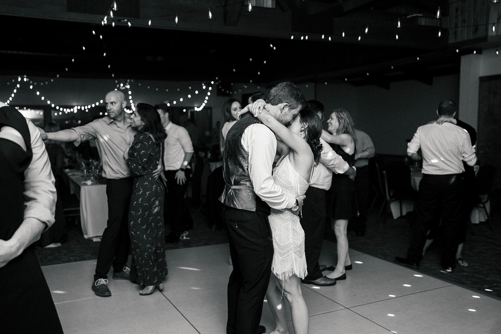 Bride in a fringed reception dress and groom embrace on a crowded dance floor under string lights, black and white.