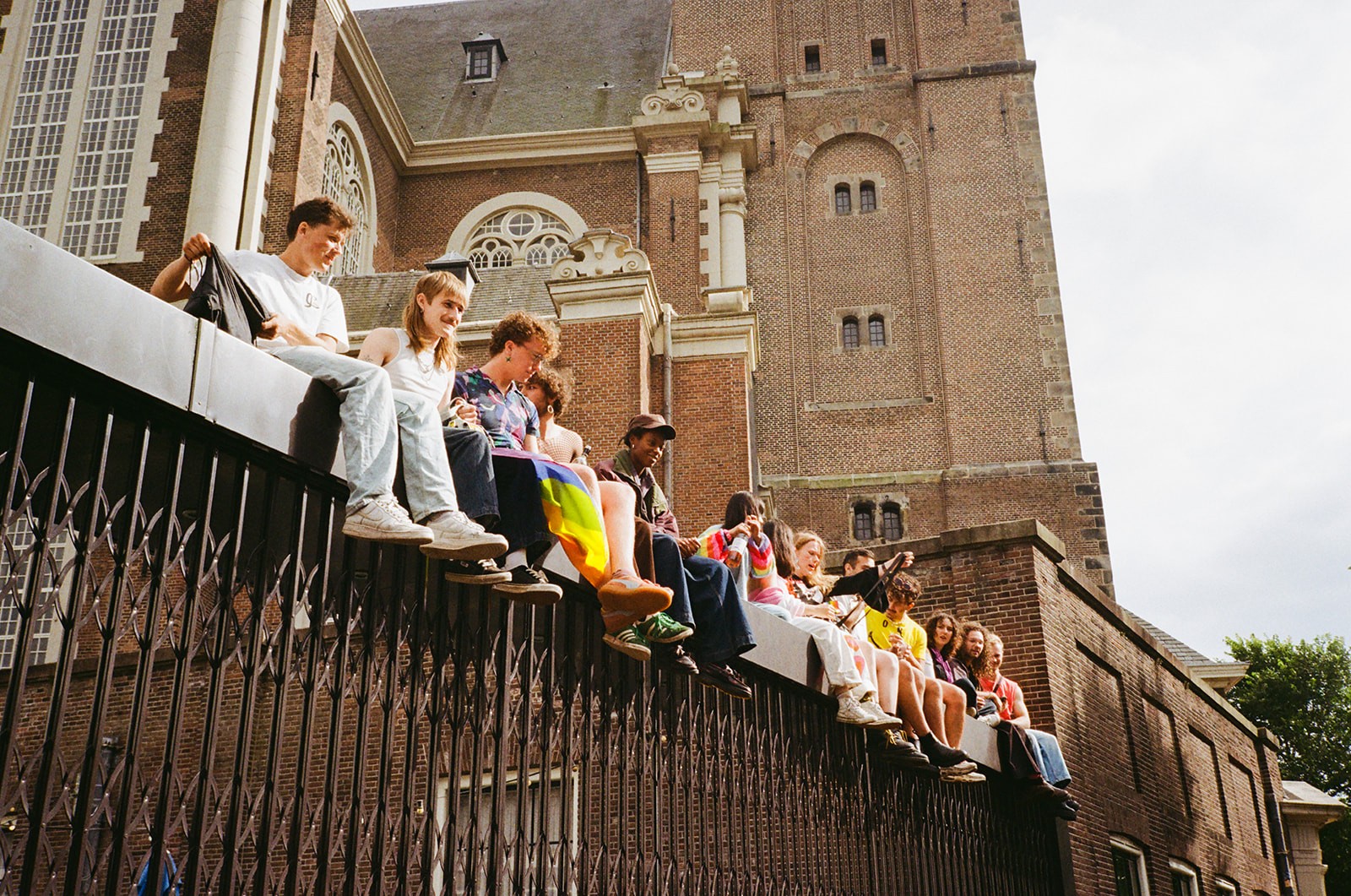Group of young people sit along a decorative iron fence outside a brick church tower in Amsterdam, chatting in the sun.