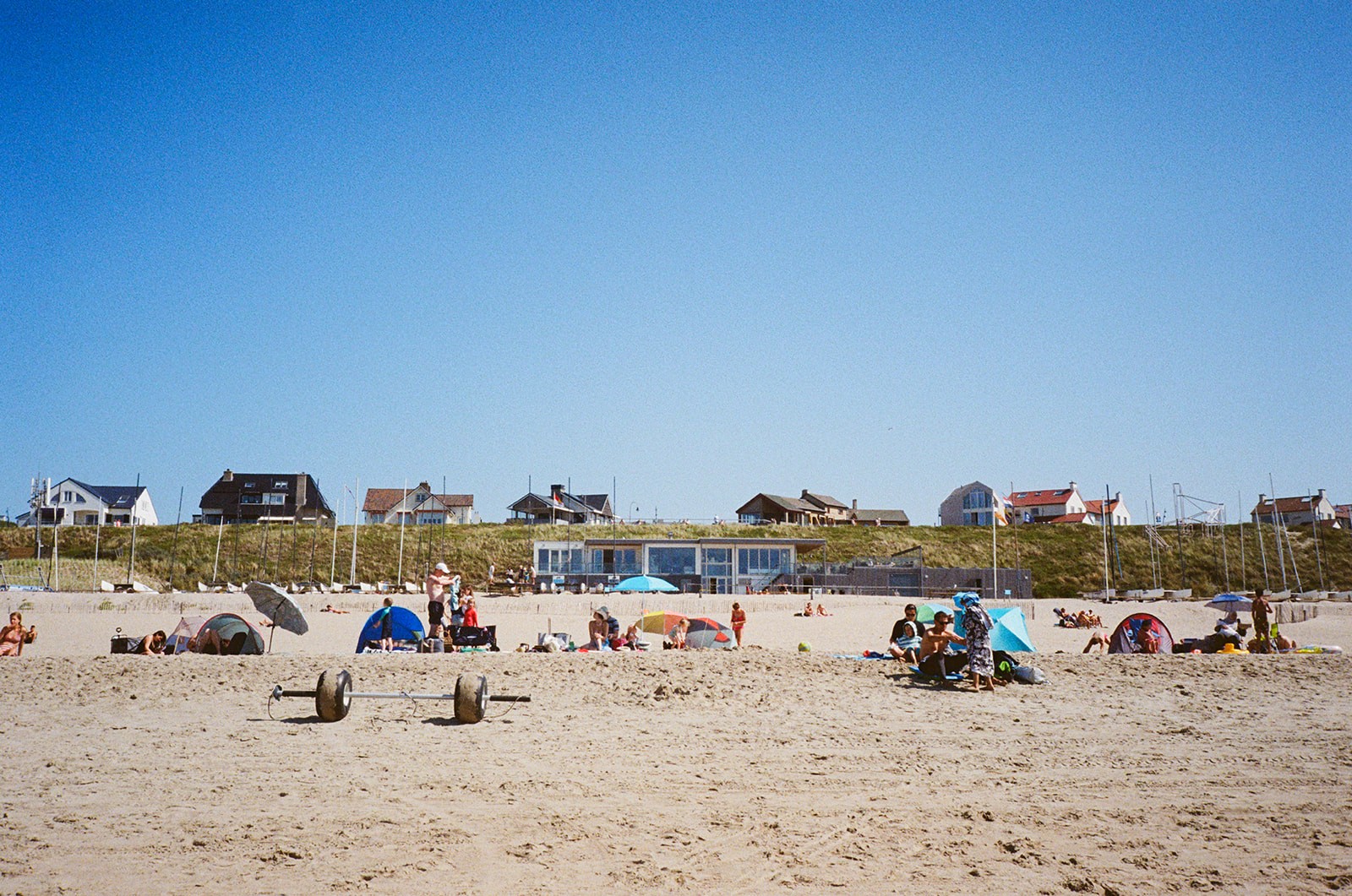 Sunbathers and beach tents dot the sand at a Dutch coastal beach club beneath dune houses and a clear sky.