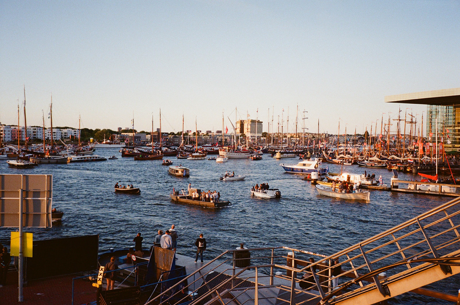 Crowds of small boats gather on Amsterdam's IJ waterway at sunset during a summer sailing festival, tall ship masts in the distance.