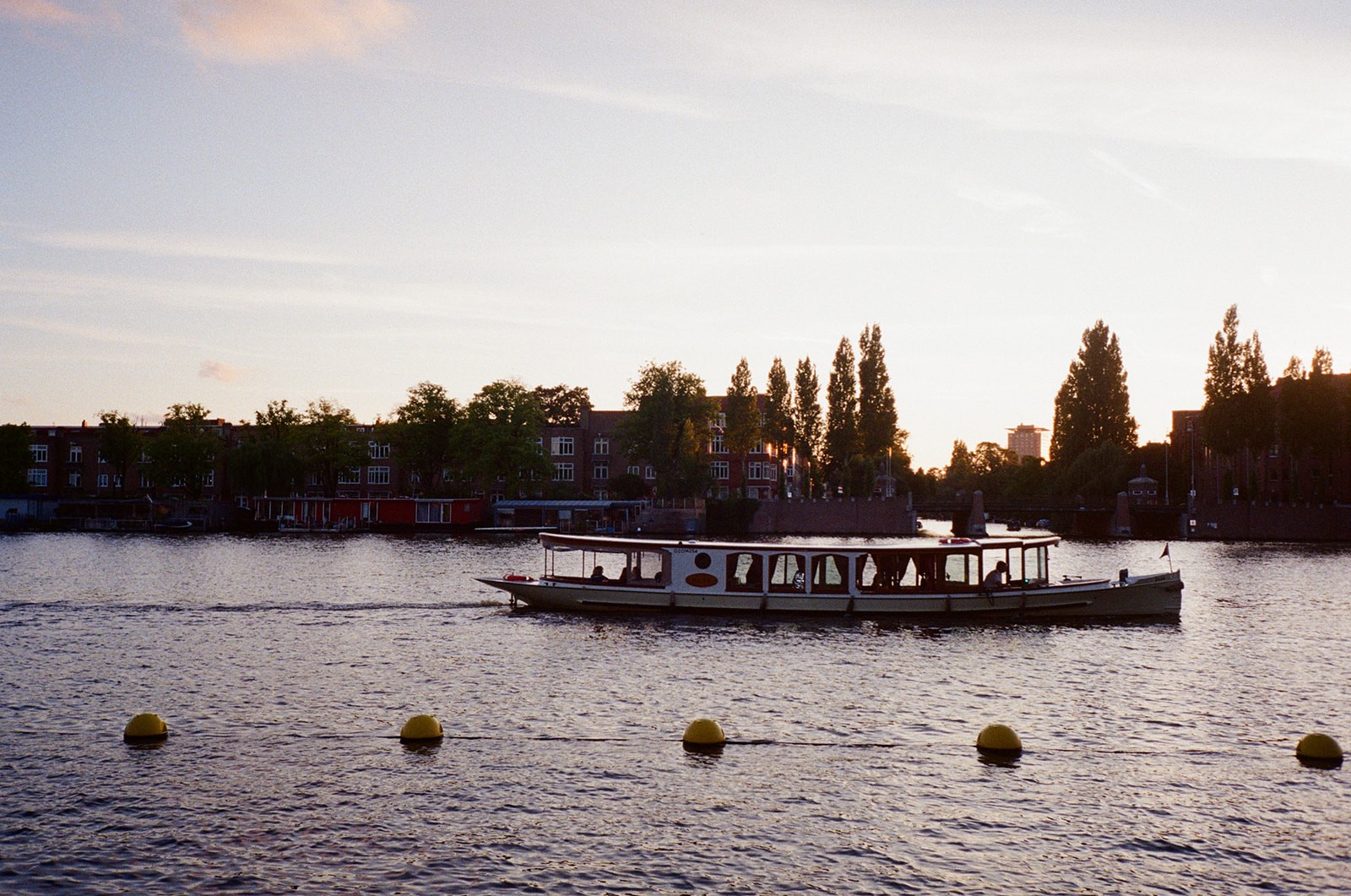 A canal tour boat glides across the Amstel river at sunset, rowhouses and trees silhouetted along the far bank.