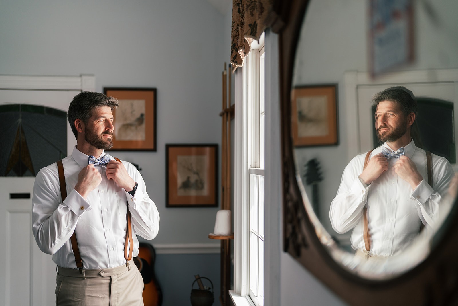 Jacob adjusting his bow tie at a window beside an oval mirror reflection