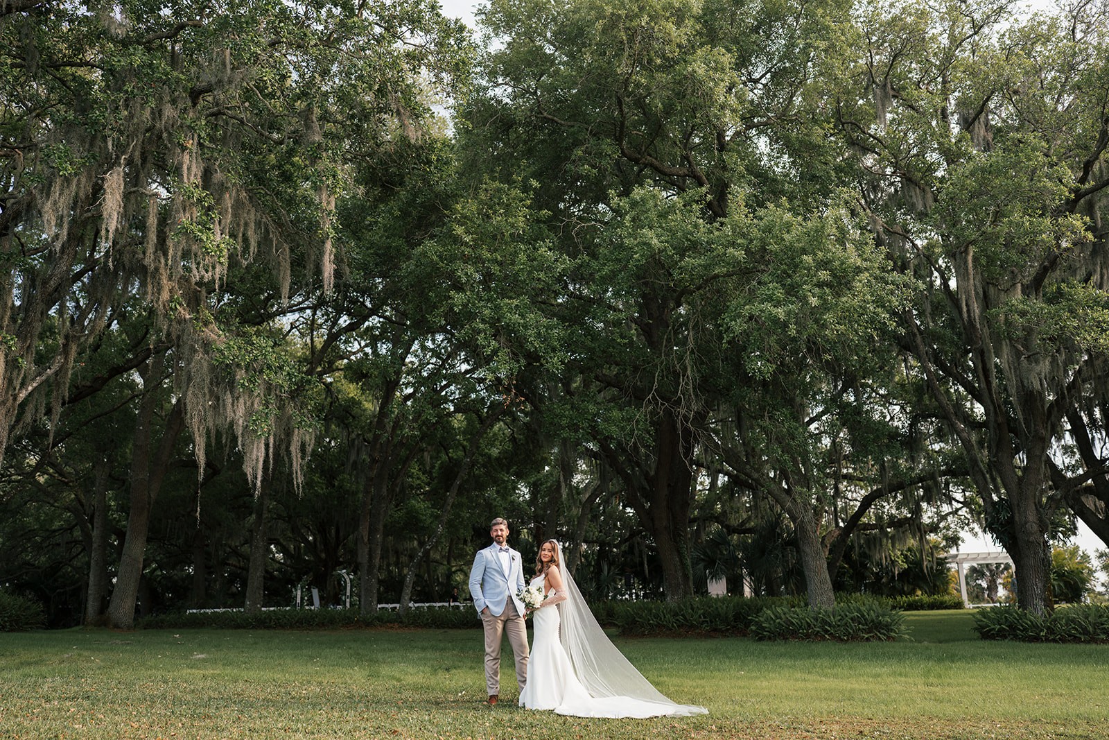 Couple stands small against sweeping Spanish moss-covered oaks on a green lawn with a white venue building in the background