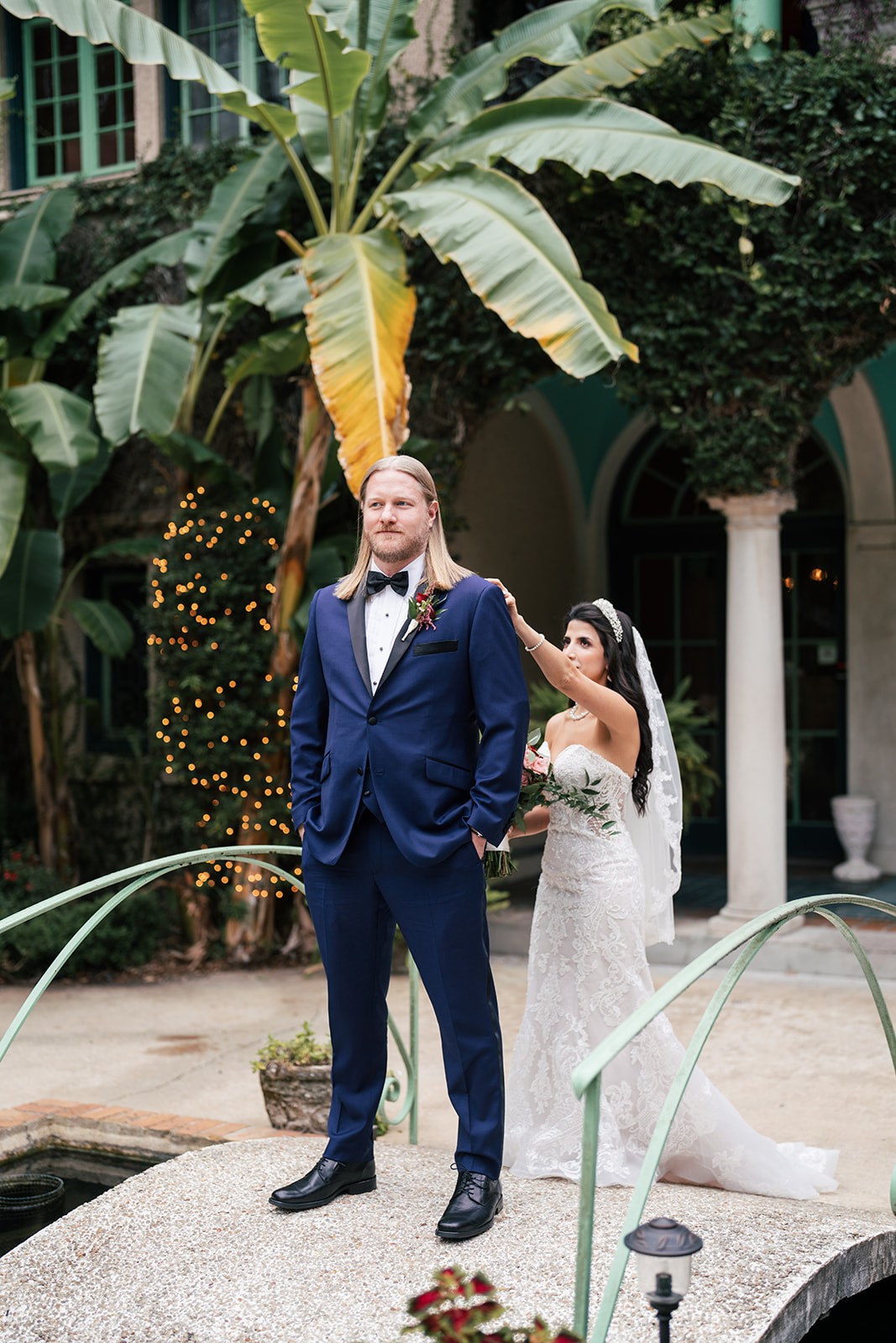 Bride touches the groom's shoulder during a first look on a courtyard footbridge amid banana palms, navy tuxedo, wedding photography