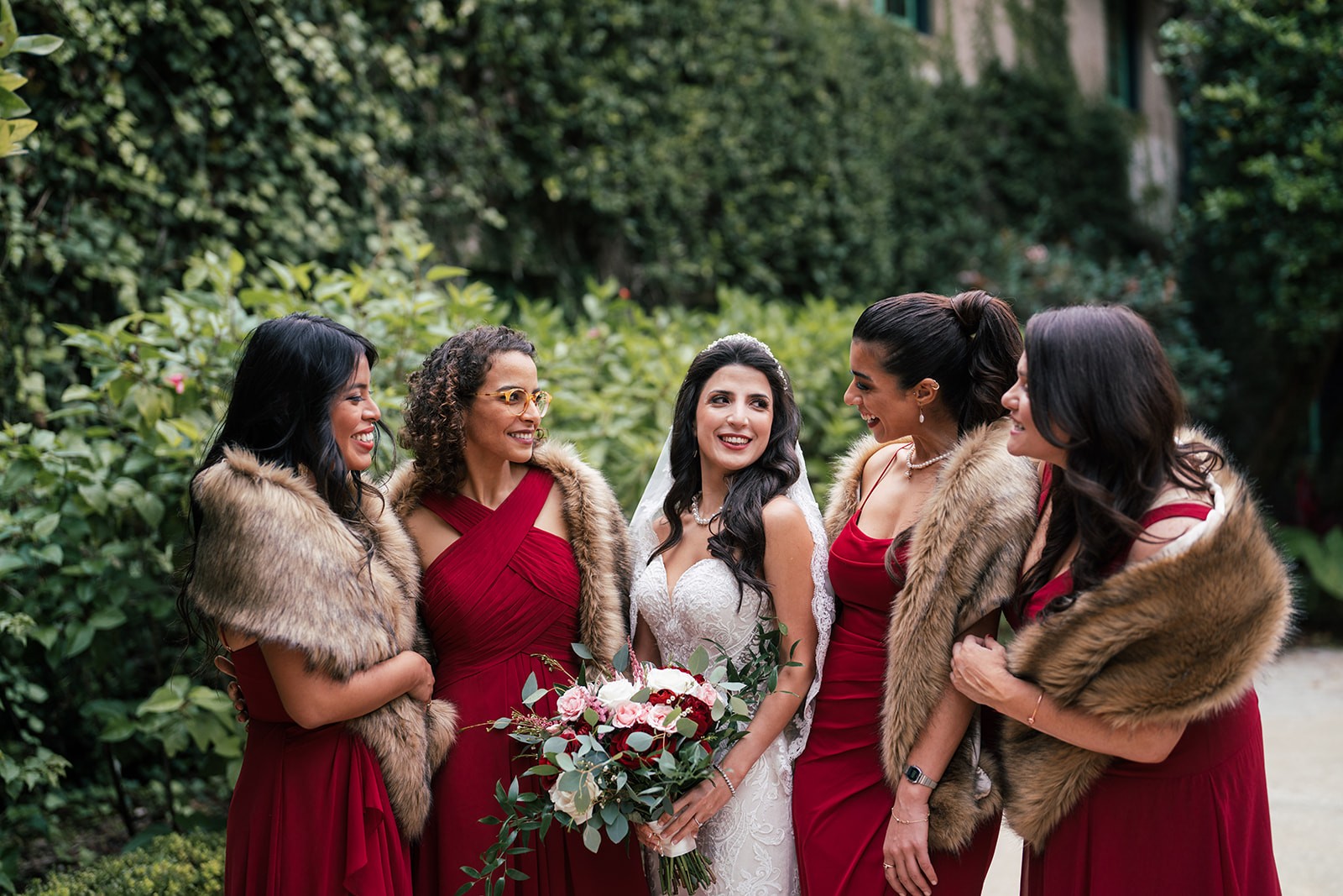 Bride laughs with bridesmaids in deep red dresses and fur stoles, holding a blush and burgundy bouquet, wedding photography