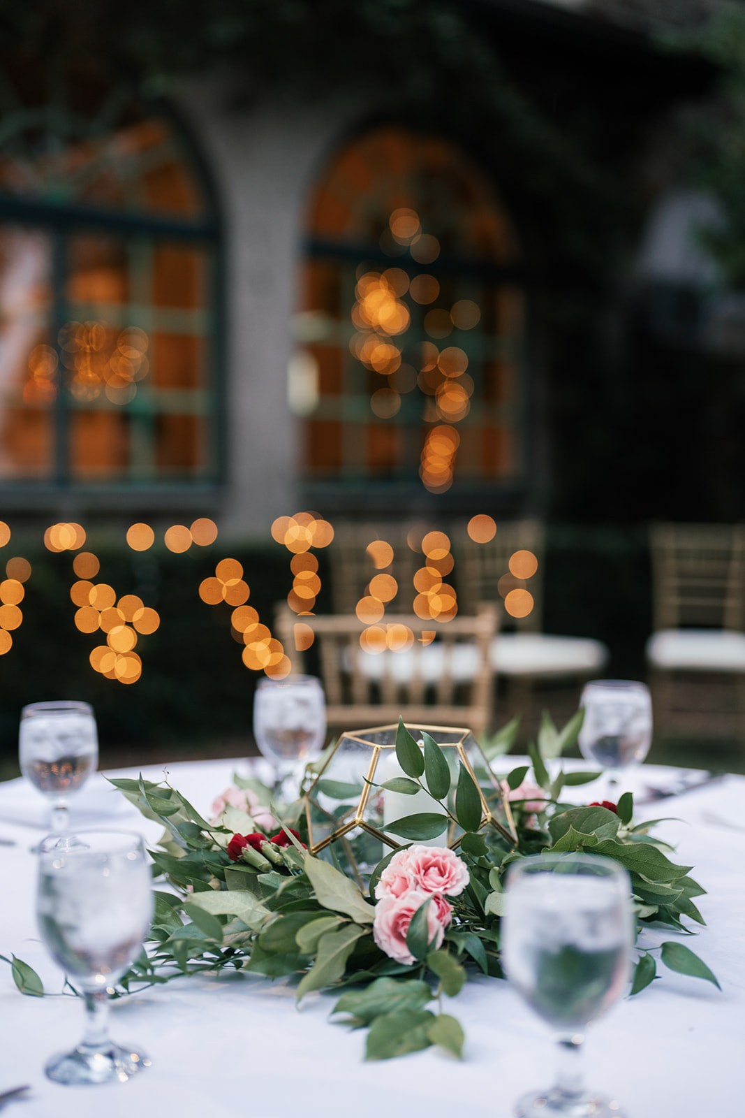 Reception centerpiece: geometric gold terrarium with pink roses and greenery, warm string lights glowing behind, wedding details
