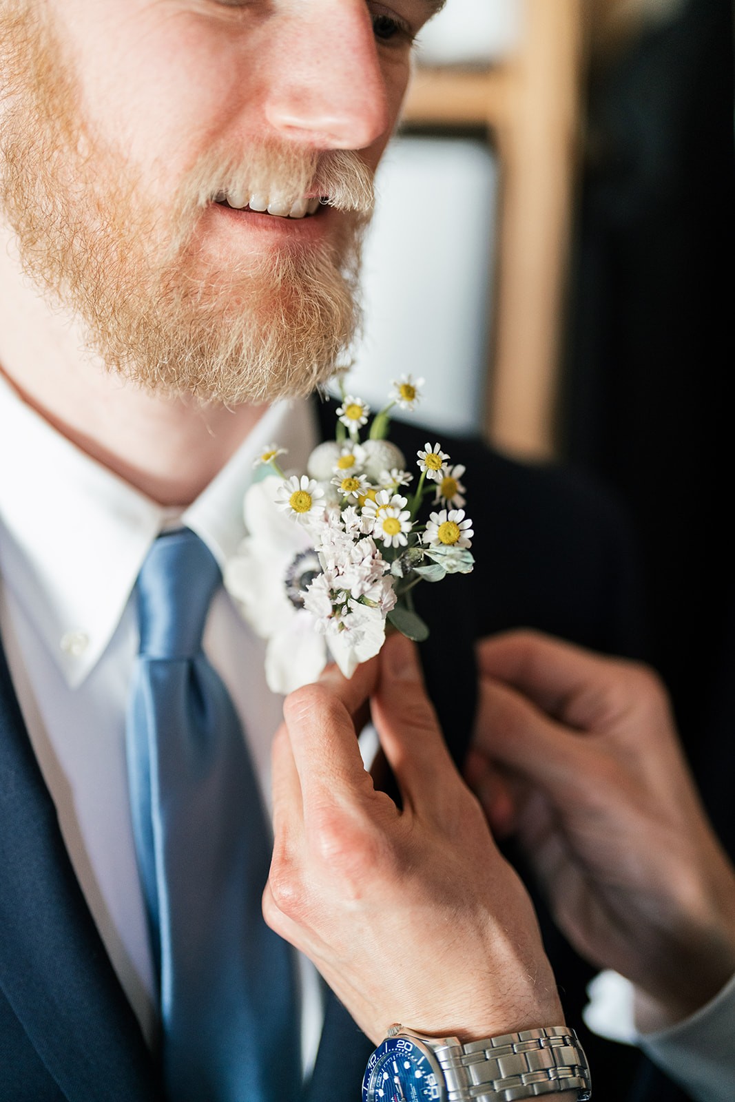 Close-up of a smiling groom having a daisy boutonniere pinned to his navy suit lapel while getting ready.