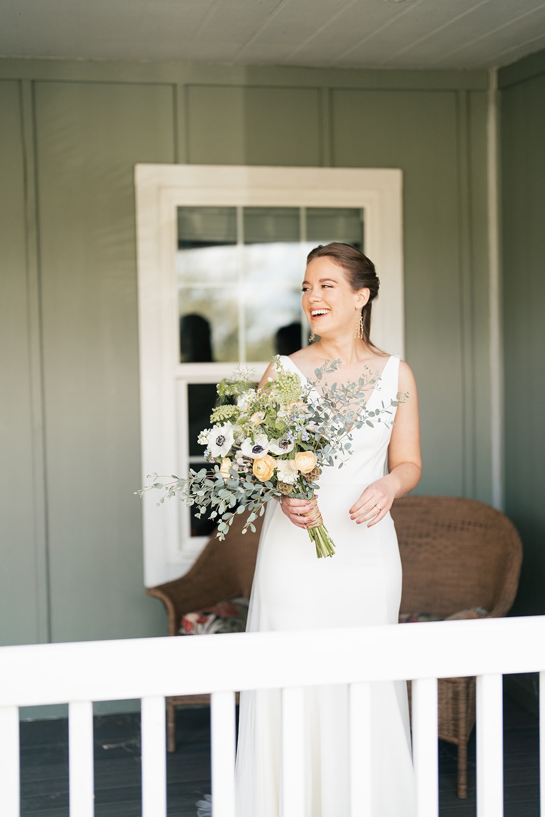 Bride in a sleeveless white gown laughing on a covered porch, holding a loose wildflower bouquet of anemones, ranunculus, and eucalyptus.