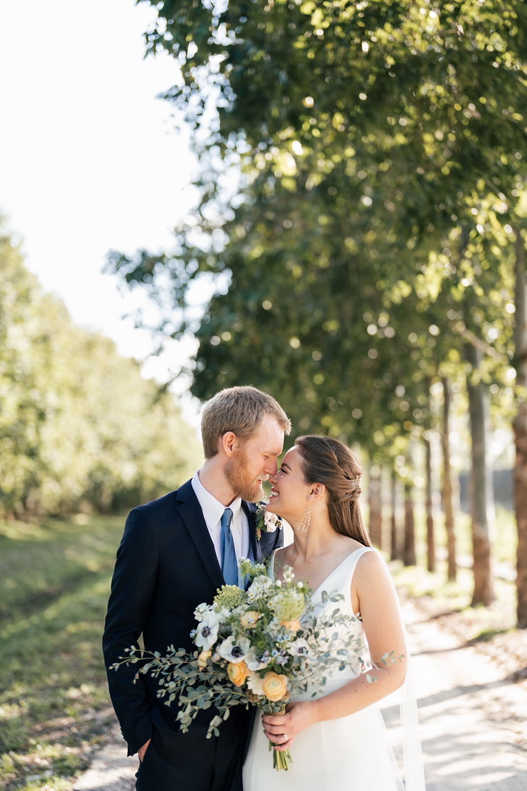 Bride and groom lean in close, nearly nose to nose and laughing, along a tree-lined path in soft afternoon light, her bouquet of anemones and ranunculus between them.